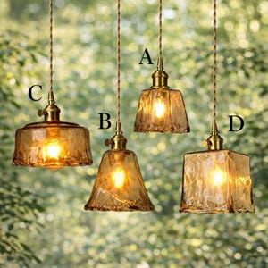 May include: Four pendant lights with amber glass shades and brass fittings hang from brown cords. The shades are various shapes: square, bell, and rounded. The lights are illuminated, casting a warm glow.