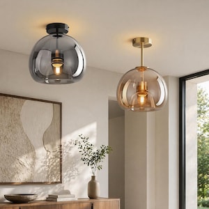 Può includere: Due moderne lampade a sospensione con paralumi in vetro. Una è di colore grigio fumé, montata a filo del soffitto con una base nera. L'altra è di colore ambra caldo, sospesa a un'asta dorata. Entrambe hanno lampadine a vista.