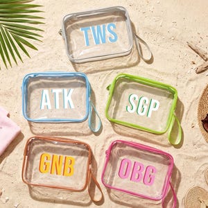 Op de afbeelding: Vijf doorzichtige, rechthoekige cosmetische tassen met verschillend gekleurde randen en initialen op een zandstrand. De tassen hebben blauwe, groene, roze, oranje en transparante randen. Een zonnehoed, zonnebril en een handdoek liggen ook op het zand.
