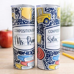 May include: Two cylindrical tumblers with a school theme. The tumblers feature a composition notebook design with a blue and white speckled background. One tumbler says "Ms. Rome" and the other says "Srta."