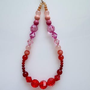 May include: A necklace featuring a gradient of beads, shifting from beige to pink and then to red. The necklace is made up of various bead shapes, including round and faceted, strung on a thin cord. The beads vary in size and texture.