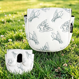 May include: Two white golf club head covers with a butterfly pattern. The covers have a white background with a repeating pattern of green butterflies. The covers are sitting on green grass.