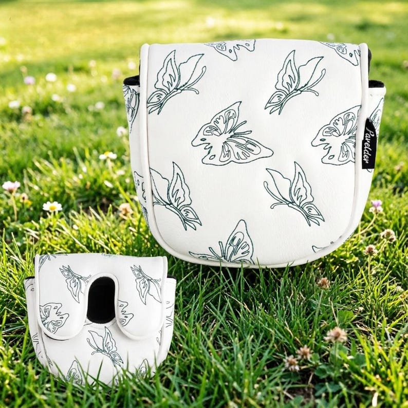 May include: Two white golf club head covers with a butterfly pattern. The covers have a white background with a repeating pattern of green butterflies. The covers are sitting on green grass.