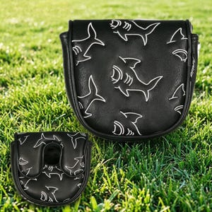 May include: Two black golf club head covers with a white shark pattern. The covers are on a green grass background. One cover is larger and rectangular, while the other is smaller and has a curved opening.