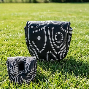 May include: Two black golf putter covers with white topographic line designs. The covers are on a green grassy surface. The larger cover has a circular design element. The smaller cover has a cutout for the putter head.