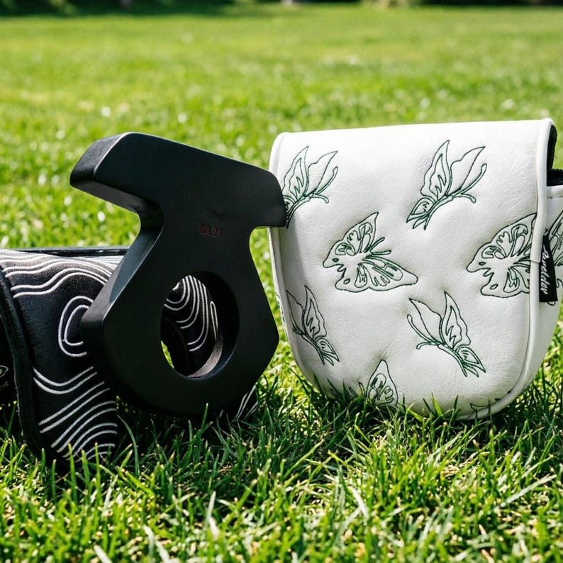 May include: A black golf putter with a unique design, next to two golf club head covers. One cover is white with green butterfly embroidery, the other is black with a white wave pattern. The items are on green grass.