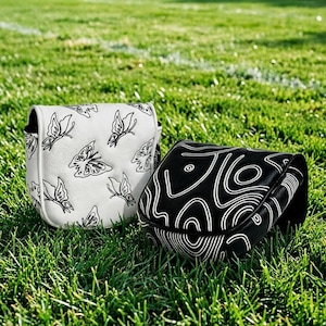 May include: Two golf club head covers rest on green grass. One is white with black butterfly designs, the other is black with a white abstract pattern. The covers are likely made of leather or a similar material.