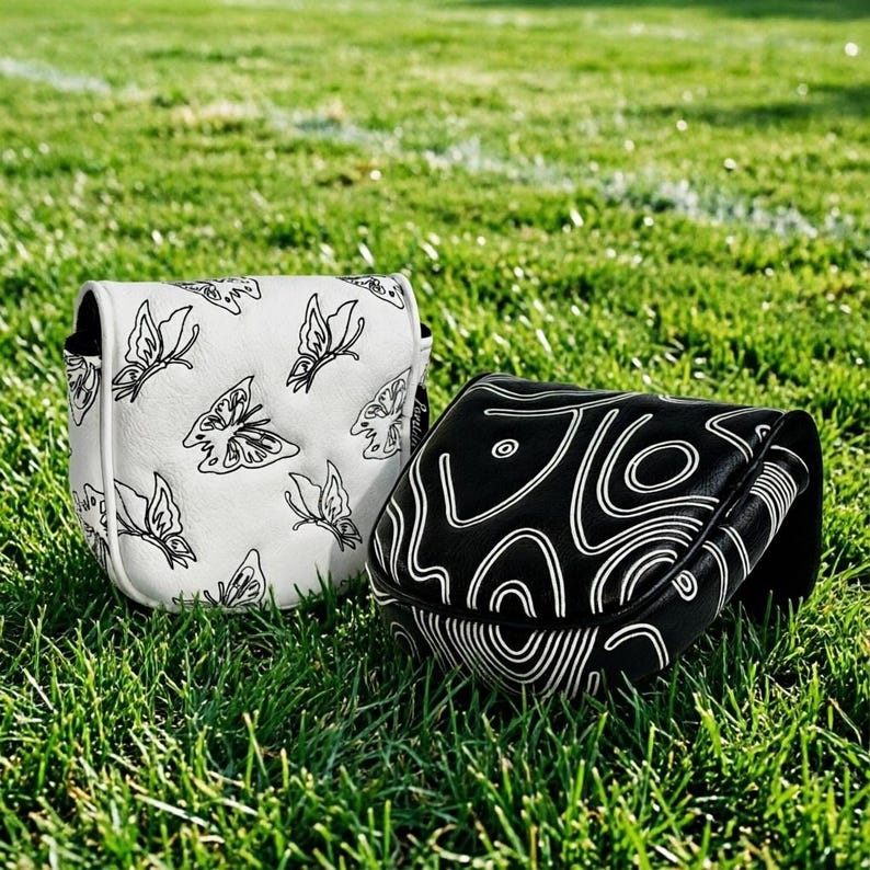 May include: Two golf club head covers rest on green grass. One is white with black butterfly designs, the other is black with a white abstract pattern. The covers are likely made of leather or a similar material.