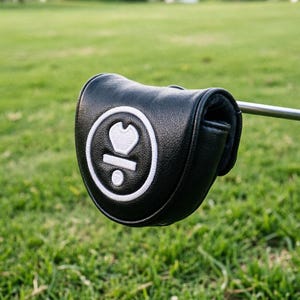 May include: A black leather golf club head cover with a white emblem featuring a heart-like shape and a dot. The cover is on a golf club, set against a backdrop of green grass.