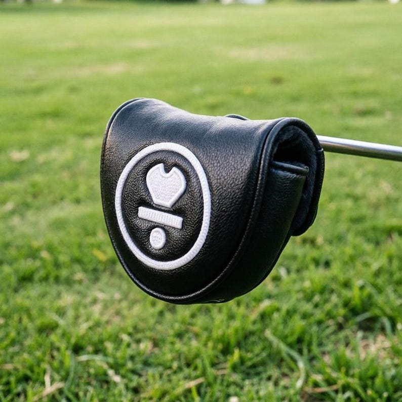 May include: A black leather golf club head cover with a white emblem featuring a heart-like shape and a dot. The cover is on a golf club, set against a backdrop of green grass.