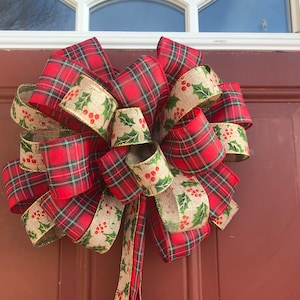 May include: A red and green plaid ribbon bow with a burlap ribbon with a holly and berry pattern. The bow is tied with a long tail and is hanging on a red door.
