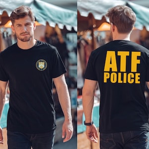 May include: A black short-sleeved t-shirt featuring a small circular emblem on the left chest and the large yellow text ATF POLICE printed on the back. The emblem includes a shield design with the words Bureau of Alcohol, Tobacco and Firearms.