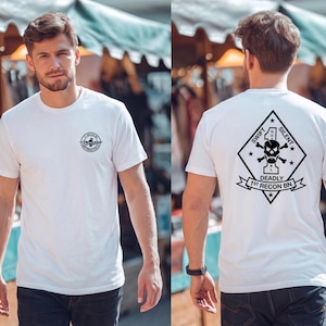 Puede incluir: Una camiseta blanca de manga corta con un pequeño logotipo circular negro en el pecho izquierdo y un gran gráfico negro en forma de diamante en la espalda. El diseño trasero incluye una calavera, tibias cruzadas y el texto Swift Silent Deadly 1st Recon Bn.