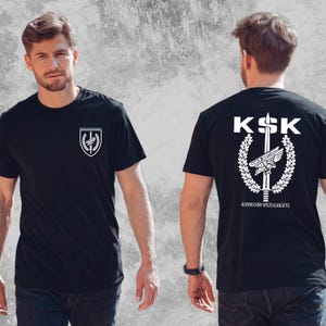 May include: A black short-sleeved t-shirt featuring a white graphic print. The front displays a small shield emblem on the left chest. The back features a large KSK logo with a sword, wings, and laurel wreath, along with the text KOMMANDO SPEZIALKRÄFTE.