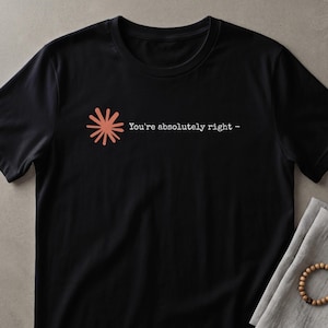 May include: A black short-sleeved t-shirt featuring a coral-colored starburst graphic on the left chest. Next to the graphic, the text "You're absolutely right -" is printed in a white, typewriter-style font across the front of the garment.