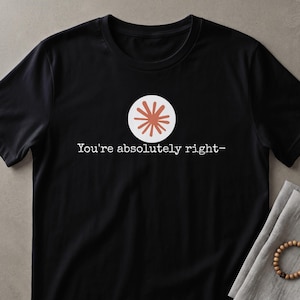 Puede incluir: Una camiseta negra de manga corta con un gráfico circular blanco que presenta un diseño de estrella de color coral en el centro. Debajo del gráfico, el texto "You're absolutely right-" está impreso en blanco con una fuente de estilo máquina de escribir.