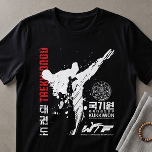 Puede incluir: Una camiseta negra de manga corta con un gran gráfico blanco de una persona realizando una patada de taekwondo. El diseño incluye el texto vertical en rojo y blanco TAEKWONDO, el logotipo circular de Kukkiwon, caracteres coreanos y el logotipo de la WTF.