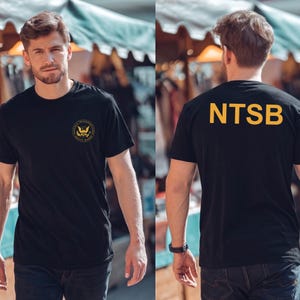 Peut inclure: Un t-shirt noir à manches courtes arborant un emblème circulaire du National Transportation Safety Board sur la poitrine gauche et le texte jaune NTSB imprimé en grandes lettres capitales sur le haut du dos du vêtement.