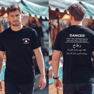 May include: A black short-sleeve t-shirt featuring a small logo on the front chest with the text Blackwater Templar Knights Baghdad Iraq. The back displays the text DANGER STAY 100 METERS BACK OR YOU WILL BE SHOT above Arabic script text.