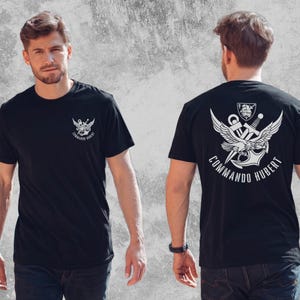 May include: A black short-sleeve t-shirt featuring a white graphic print. The front displays a small emblem with an anchor, wings, and a sword. The back features a larger version of the same emblem with the text COMMANDO HUBERT printed below.
