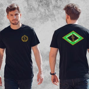 May include: A black t-shirt featuring a circular gold logo on the front left chest with the text Jungle Warfare Training Center. The back displays a diamond-shaped graphic with green and yellow borders, a central snake, and text including United States Marine Corps.