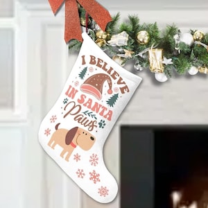 May include: A white Christmas stocking with the text "I Believe in Santa Paws" and a cartoon dog illustration. The stocking is decorated with snowflakes and a Santa hat. A garland with ornaments and red bows is above the fireplace.