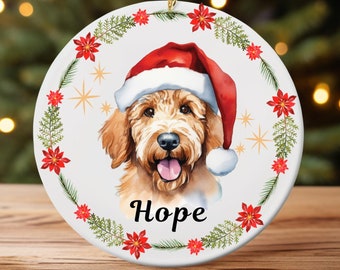 May include: A round, white Christmas ornament featuring a watercolor illustration of a golden doodle wearing a Santa hat. The ornament is encircled by a wreath of red poinsettias and green pine needles, with the word "Hope" below the dog's face.
