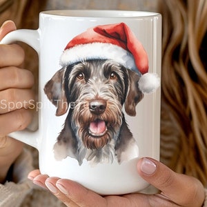 May include: White ceramic mug featuring a watercolor illustration of a dog wearing a Santa hat. The dog has brown and gray fur and a happy expression. The mug has a large handle and is held by a person.