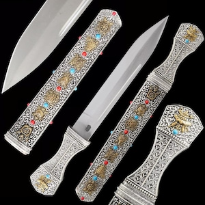 Könnte beinhalten: Ein verzierter Dolch mit silberner Klinge und Griff. Der Griff und die Scheide sind silberfarben mit goldenen Akzenten, Türkis und roten Steinen. Das Design weist komplizierte Muster und Symbole auf.