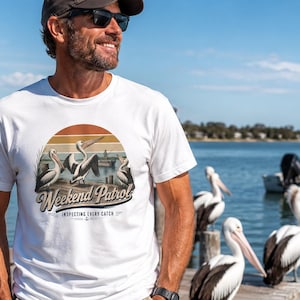 Może przedstawiać: Biały t-shirt z krótkim rękawem z okrągłą grafiką przedstawiającą zachód słońca, trzy pelikany oraz napis "Weekend Patrol" nad "INSPECTING EVERY CATCH" z małą ikoną kotwicy wyśrodkowaną pod tekstem na przodzie odzieży.