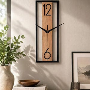 Op de afbeelding: Een rechthoekige wandklok met een zwart frame en een licht houten wijzerplaat. De klok heeft zwarte wijzers en grote zwarte cijfers op de posities 12 en 6. De klok is aan een lichtgekleurde muur bevestigd.