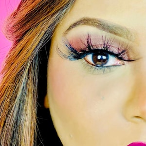 May include: Close-up of an eye with long, dark eyelashes and pink eyeshadow. The eye is brown, and the skin has a smooth texture. The hair is a mix of brown and blonde, and the background is a bright pink.
