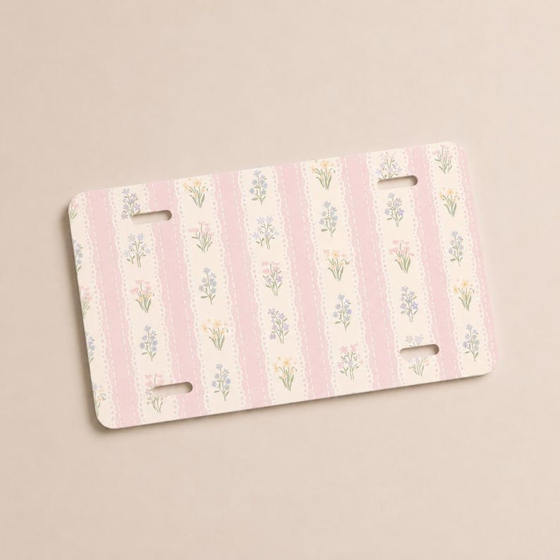 Op de afbeelding: Rechthoekige nummerplaat met een bloemenpatroon. Het ontwerp bevat verticale roze strepen met een kantachtige rand, afgewisseld met kleine bloemenillustraties in pastelkleuren. De plaat heeft vier montagegaten.