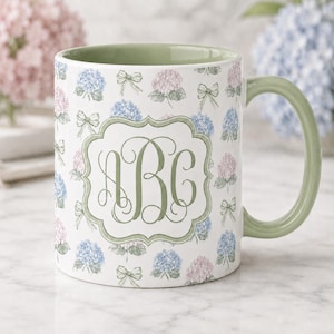 May include: White ceramic mug with a green handle and interior. The mug is decorated with a pattern of blue and pink hydrangea flowers and green bows. A green monogram with the letters "ABC" is centered on the mug.
