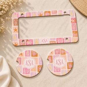 May include: A license plate frame and two car coasters with a beach theme. The items feature a repeating pattern of seashells, starfish, and coral in pink, orange, and yellow squares. Each item has the initials "KSA" in a white oval.