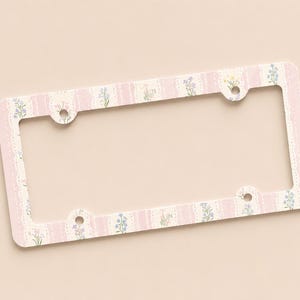 Op de afbeelding: Een rechthoekig kentekenplaatraam met een bloemenontwerp. Het frame heeft een roze en cr&egrave;mekleurig kleurenschema met een herhalend patroon van verticale strepen en kleine bloemen. Het frame heeft vier montagegaten.