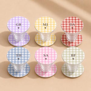 May include: Six circular phone grips with a gingham pattern in purple, yellow, red, blue, pink, and green. Each grip features two black capital letters and a small black heart icon centered on the top surface, with a clear, ribbed plastic base.