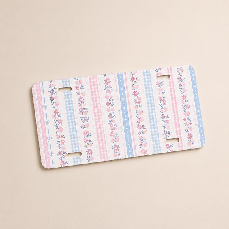 Op de afbeelding: Een rechthoekige nummerplaat met een bloemen- en ruitpatroon in pastelroze en blauw. Het ontwerp bevat verticale strepen van bloemen, stippen en ruitjes, met kleine strikjes. De plaat heeft vier sleuven voor montage.