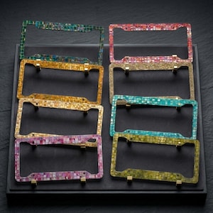 May include: A collection of mosaic-tiled license plate frames in various colors, including green, gold, pink, and blue. Each frame is rectangular with rounded corners and features a mosaic pattern. The frames are displayed on a black surface.