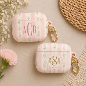 May include: Two pink and white striped AirPod cases with floral accents and gold-colored carabiner clips. One case has the initials "ACB" in pink, and the other has "KSR" in gold.
