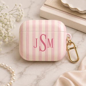 May include: Pink and white striped AirPod case with the initials "JSM" in a matching pink font. The case has a gold-colored clasp and is set against a marble surface. The overall aesthetic is feminine and stylish.