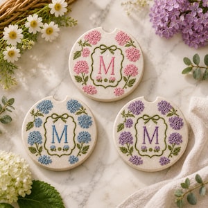 May include: Three round car coasters with floral designs. Each coaster features a different color of hydrangea flowers: pink, blue, and purple. The center of each coaster has the letter "M" in a decorative frame.