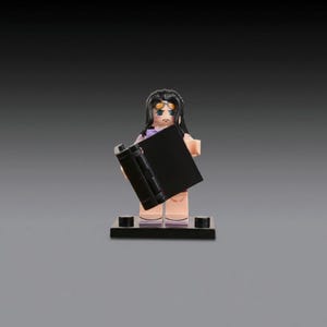 May include: A Lego figurine of a character with black hair, wearing a purple top and holding a black book. The figurine has a sad expression and is standing on a black base.