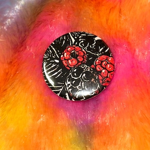 May include: A circular pin-back button featuring a high-contrast graphic illustration of several raspberries. The design uses a black background with white line-art details and vibrant red coloring for the berries, creating a bold visual effect.