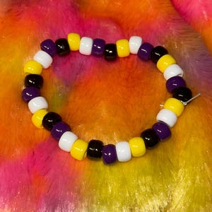 Könnte beinhalten: Ein kreisförmiges Armband aus abwechselnden Kunststoffperlen in den einfarbigen Tönen Gelb, Weiß, Violett und Schwarz. Die Perlen sind auf einer dünnen, flexiblen Schnur aufgereiht und bilden ein sich wiederholendes Muster dieser vier Farben entlang des gesamten Rings.
