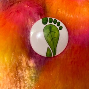 May include: A circular pinback button featuring a white background with a green graphic illustration of a footprint shaped like a leaf. The footprint design includes a large leaf-shaped sole and four smaller circular leaf-shaped toes at the top.