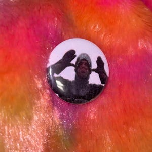 May include: A circular pinback button featuring a graphic print of a person wearing a dark hooded garment and gloves, with their hands raised near their face. The button has a white background and a glossy finish, displaying a clear photographic image.