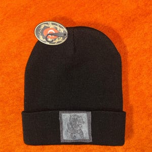May include: Black knit beanie with a folded brim. A rectangular patch with a tiger illustration is sewn on the brim. A tag with a circular design is attached to the beanie.