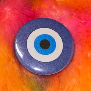 May include: A circular pin-back button featuring a classic evil eye design. The graphic consists of a dark blue outer ring, a white middle ring, a bright blue inner circle, and a solid black center pupil. The surface has a smooth, glossy finish.