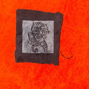 May include: A square fabric patch with a dark brown border and a light gray center panel. The center panel features a dark gray illustration of a tiger in a crouching position. The edges of the fabric patch are frayed and unevenly cut.